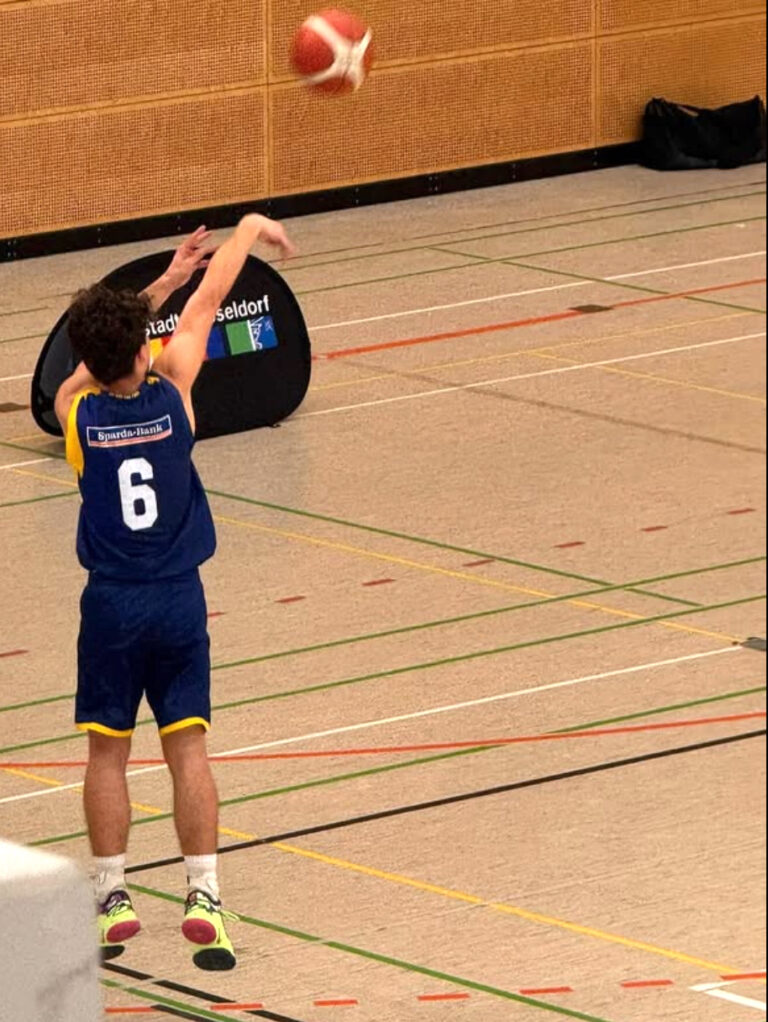 Basketballspieler beim Wurf, fokussiert, blaue Uniform mit gelben Akzenten, Halle.