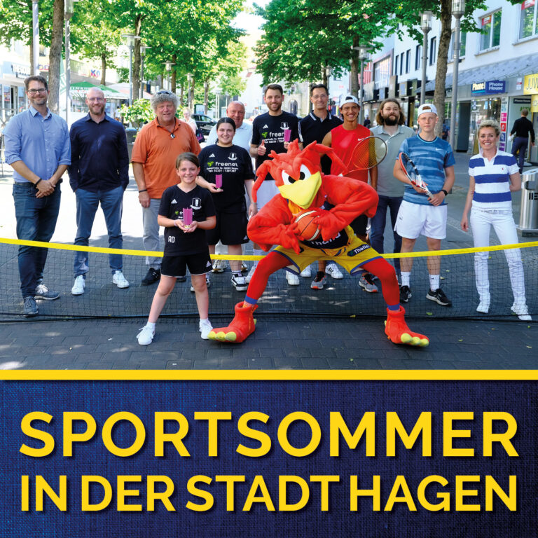 Eine Gruppe von Menschen, einige lächeln, mit einem roten Maskottchen vor einem Tennisschläger.