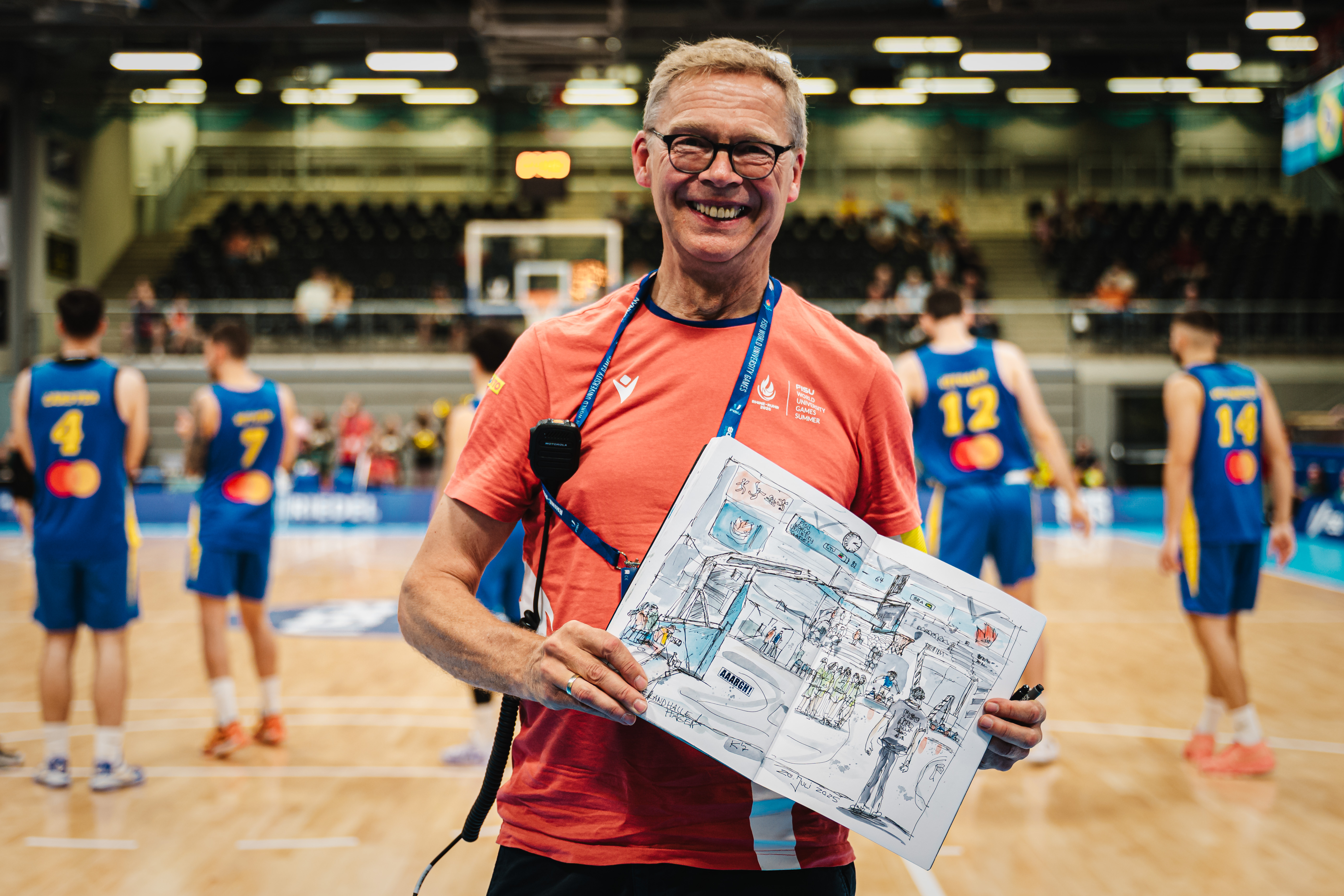 Ein lächelnder Mann in rotem Shirt hält ein kunstvolles Bild in einer Sporthalle.