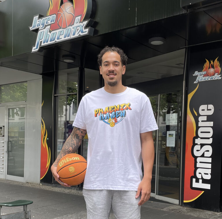 Mann mit Lächeln, trägt weißes Phoenix-Shirt, hält Basketball, steht vor Gebäude mit Phoenix-Logo.