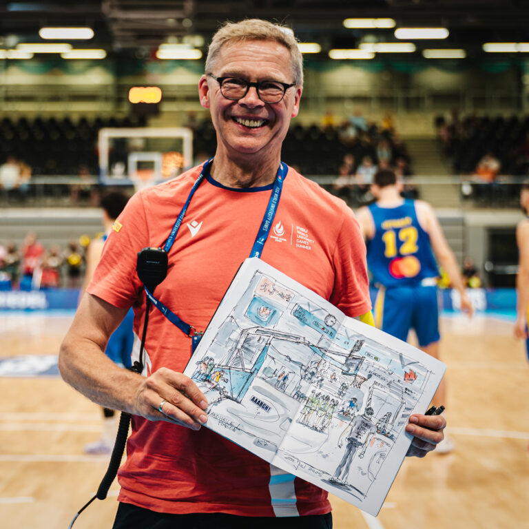 Älterer Herr lächelt und hält Zeichnung in Sporthalle, trägt oranges Shirt.