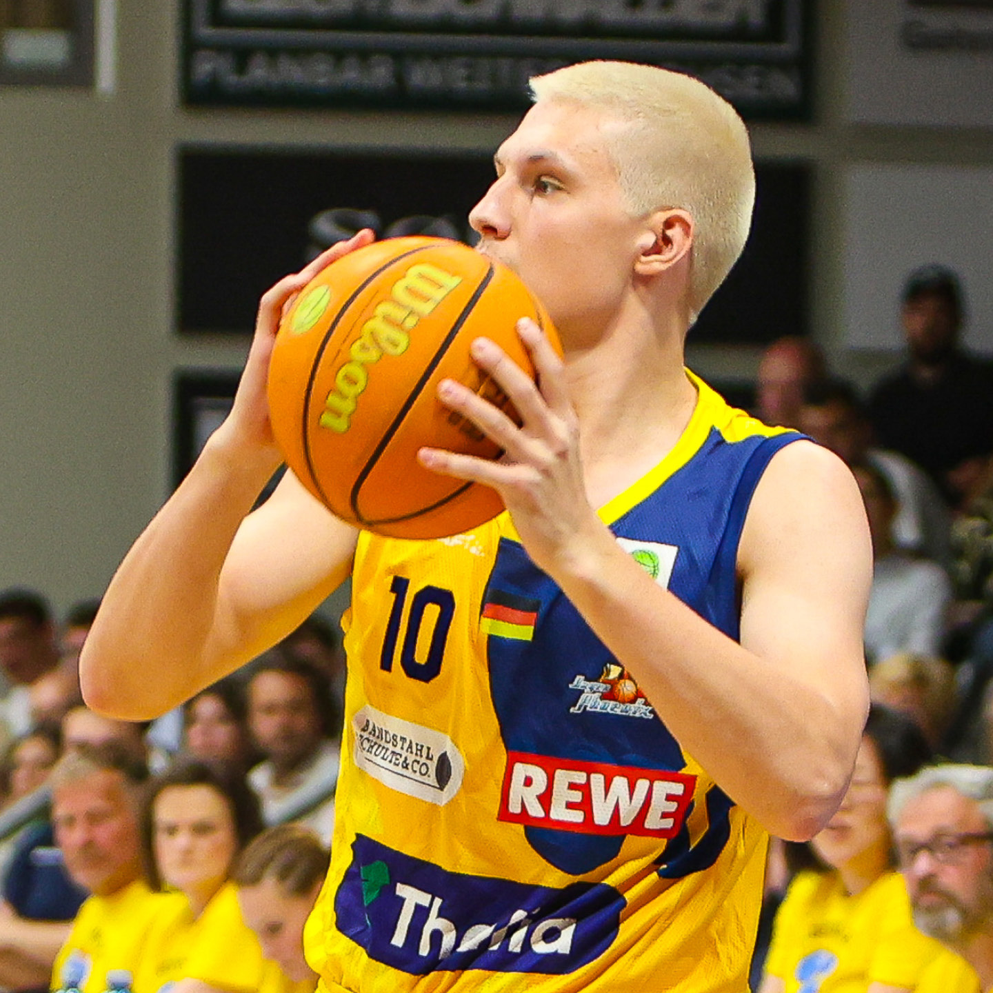 Ein Basketballspieler mit blondem Haar konzentriert sich, hält einen orangefarbenen Ball in gelbem Trikot.