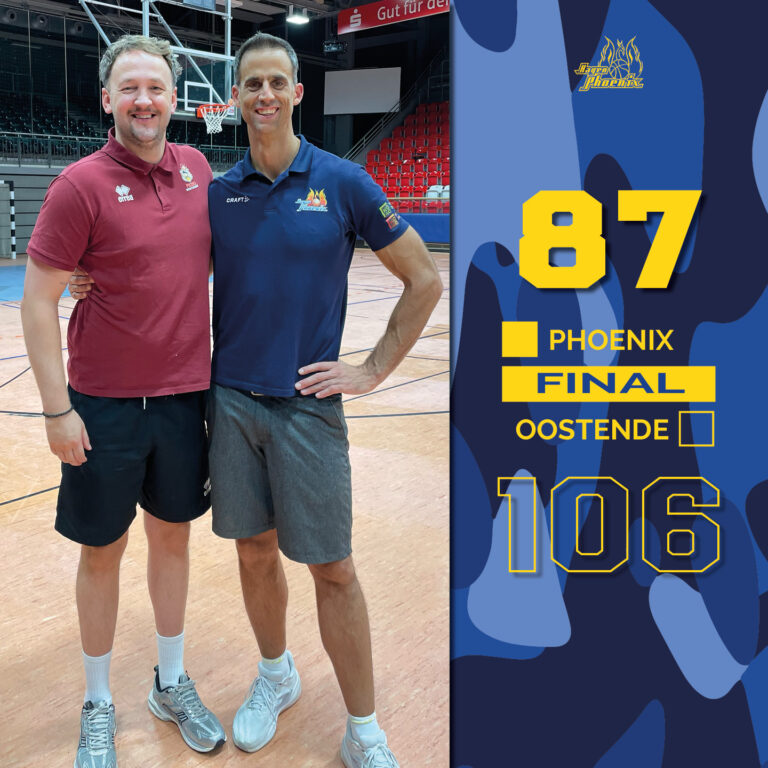 Zwei Männer lächeln fröhlich in einer Sporthalle mit einem Basketballplatz.