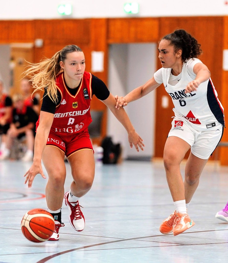 Zwei Basketballspielerinnen im Spiel, konzentriert, tragen rote und weiße Trikots.