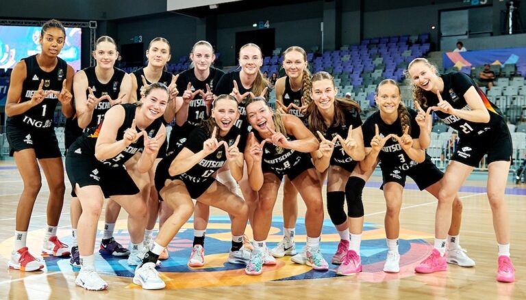 Eine fröhliche Frauen-Basketballmannschaft posiert auf dem Spielfeld in schwarzen Trikots.