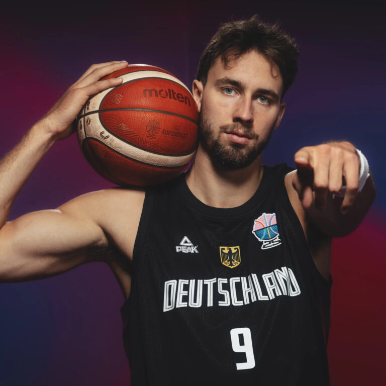 Ein Basketballspieler im Deutschland-Trikot hält einen Ball und zeigt entschlossen.