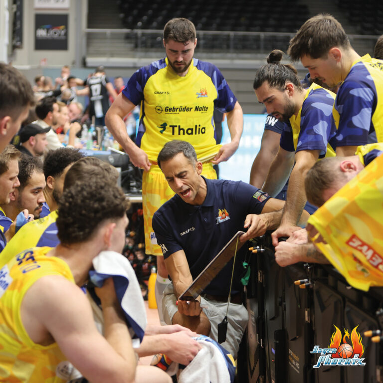 Ein Trainer in einem blauen Shirt spricht motiviert mit Basketballspielern in gelben Trikots auf einer Bank.