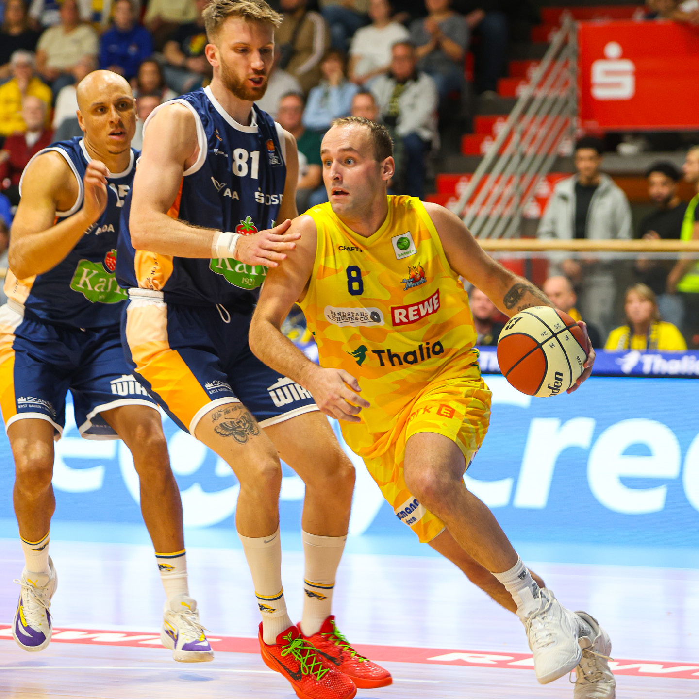 Ein Basketballspieler im gelben Trikot dribbelt dynamisch, verfolgt von gegnerischen Spielern in blauen Trikots.