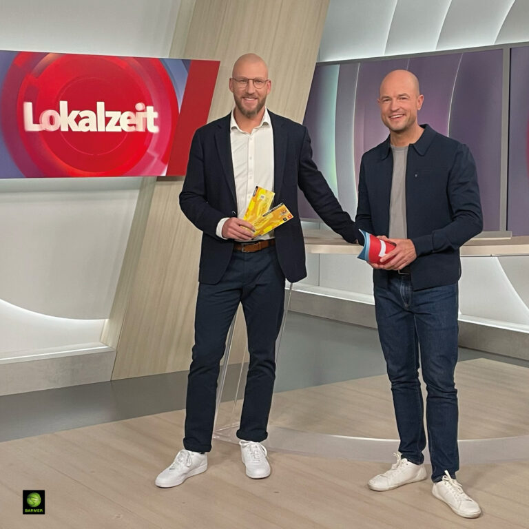 Zwei Männer lächeln in einem Studio, beide in formeller Kleidung mit Sportschuhen.