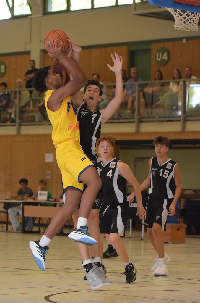 Ein junger Mann im gelben Basketball-Trikot springt zum Korb, während ein Gegenspieler ihn im schwarzen Trikot blockiert.