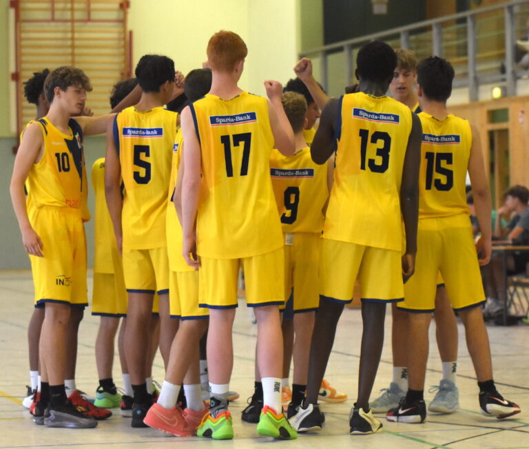 Ein Jugendbasketballteam hebt die Fäuste, alle tragen gelbe Trikots mit Nummern