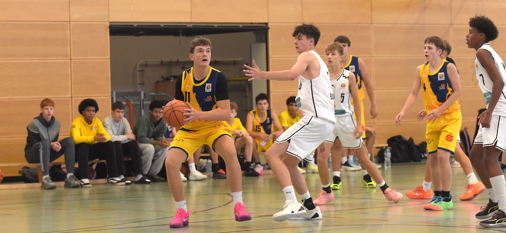 Basketballspieler in gelbem Trikot blickt konzentriert auf den Korb, während Gegenspieler in weißer Kleidung verteidigt; Holzwand und Team im Hintergrund