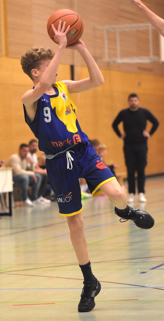 Ein junger Basketballspieler springt und wirft, mit einem fokussierten Ausdruck und einem gelb-blauen Trikot.