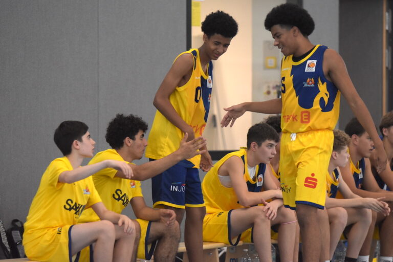 Eine Gruppe junger Basketballspieler in gelben Trikots auf der Bank, fröhlich.