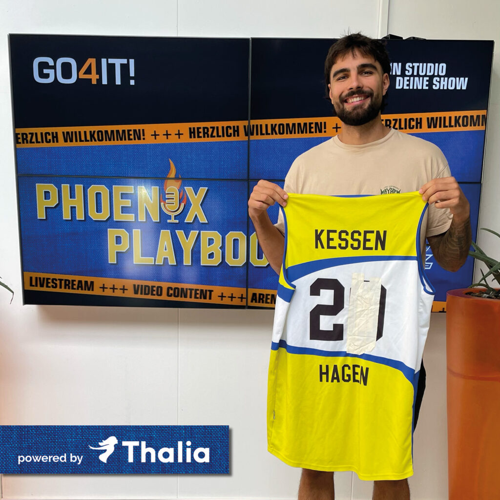 Junger Mann lächelt und hält gelbes Basketballtrikot, Tafeln im Hintergrund.