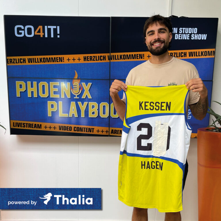 Junger Mann lächelt und hält gelbes Basketballtrikot, Tafeln im Hintergrund.