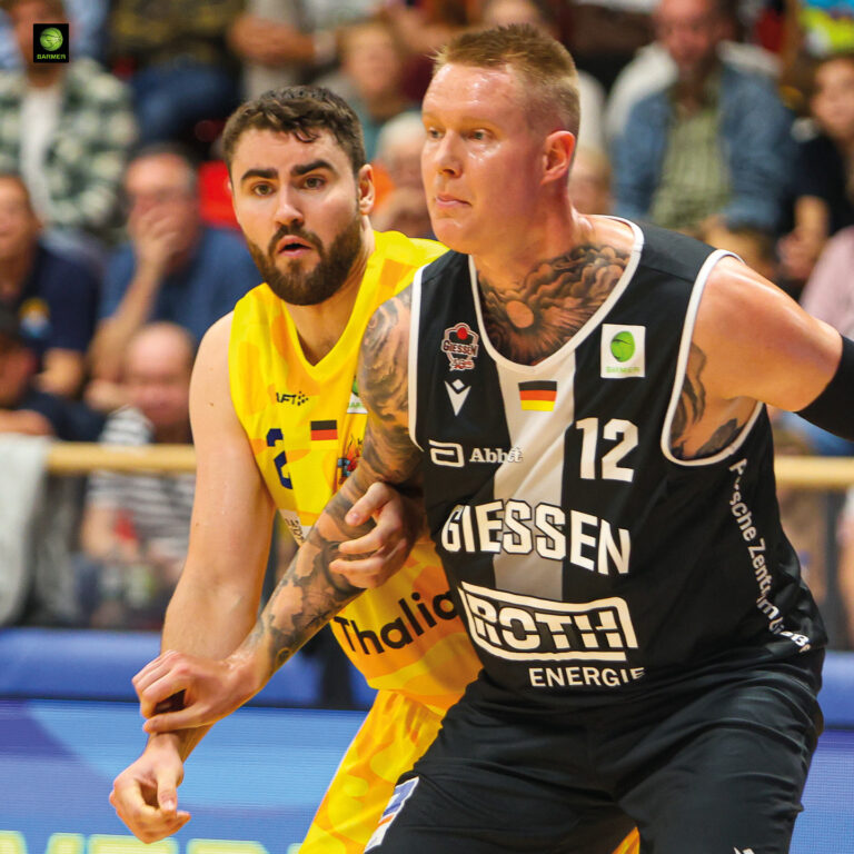 Zwei Basketballspieler in Aktion, ein Spieler in schwarz, tätowiert, entschlossen.