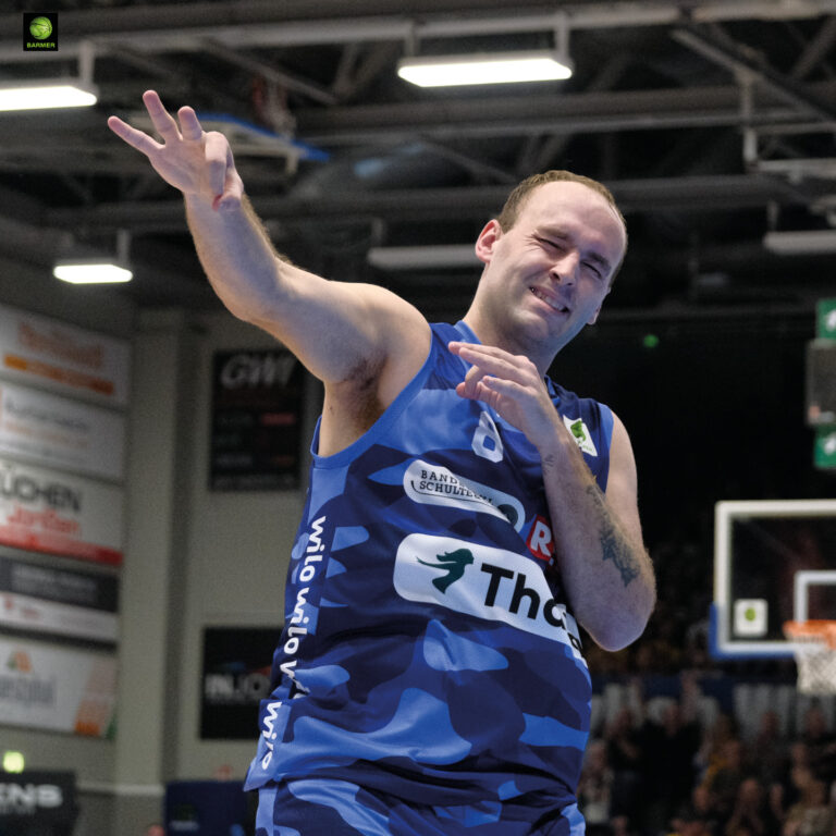 Ein Basketballspieler in blauem Trikot posiert mit humorvollem Ausdruck.