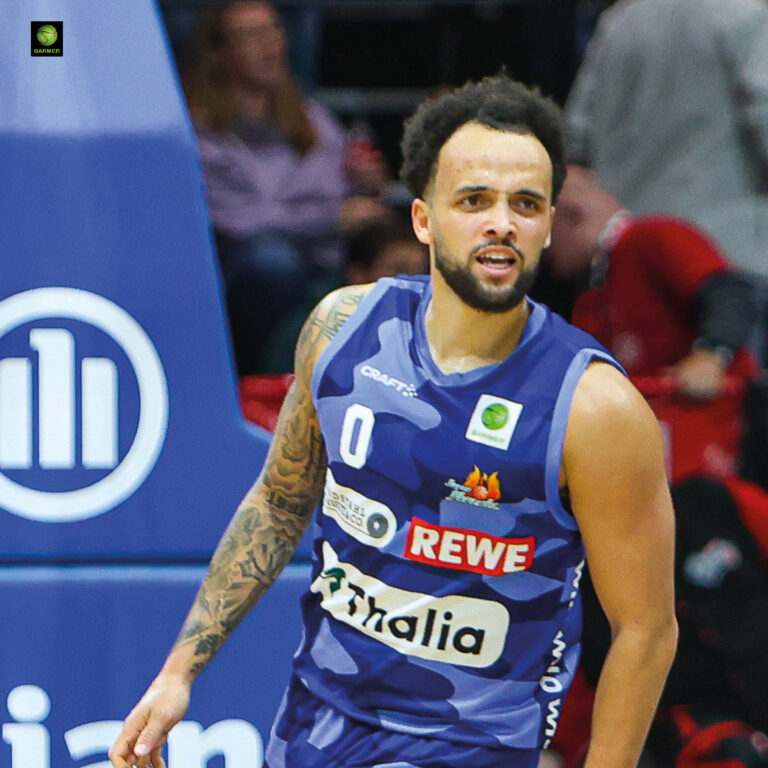 Ein junger Basketballspieler mit Bart und lockigem Haar wirkt fokussiert im blauen Trikot