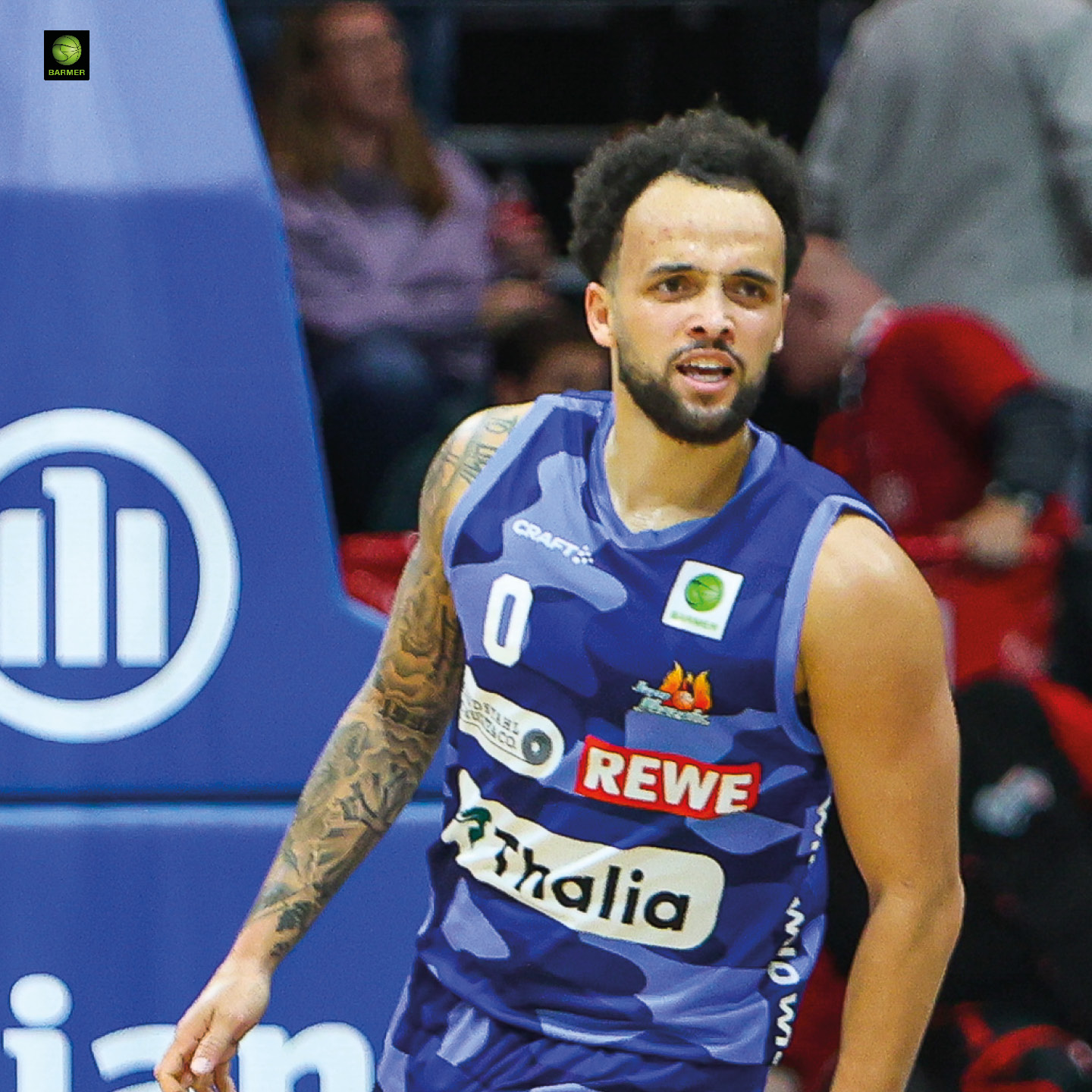 Ein junger Basketballspieler mit Bart und lockigem Haar wirkt fokussiert im blauen Trikot