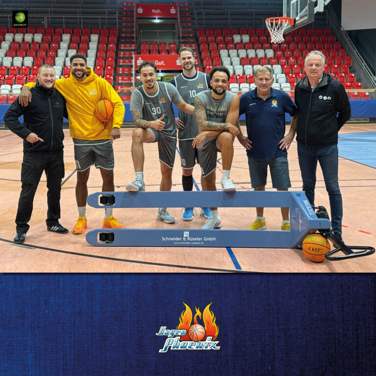 Sechs Männer stehen lachend in einer Sporthalle mit rotem Sitzbereich und Basketballkorb