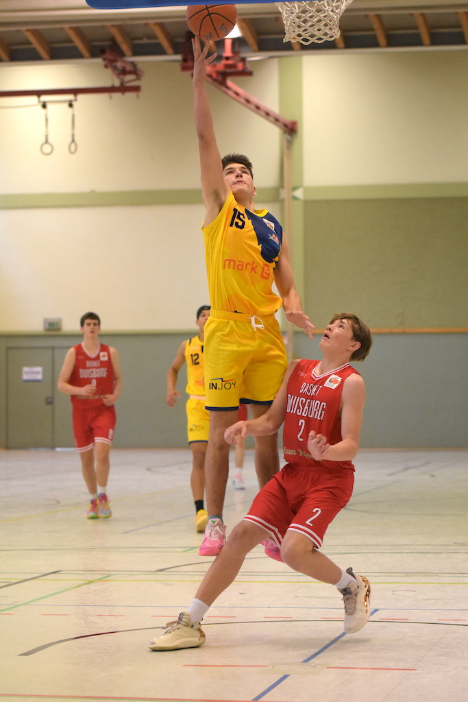 Ein Basketballspieler in gelbem Trikot springt zum Korb, während ein Gegner in Rot versucht zu blocken.