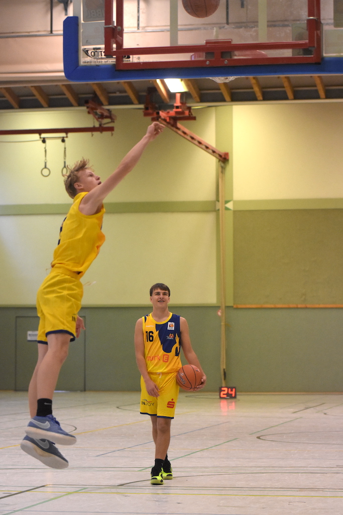 Zwei junge Männer in gelben Basketballuniformen in einer Sporthalle, einer springt.