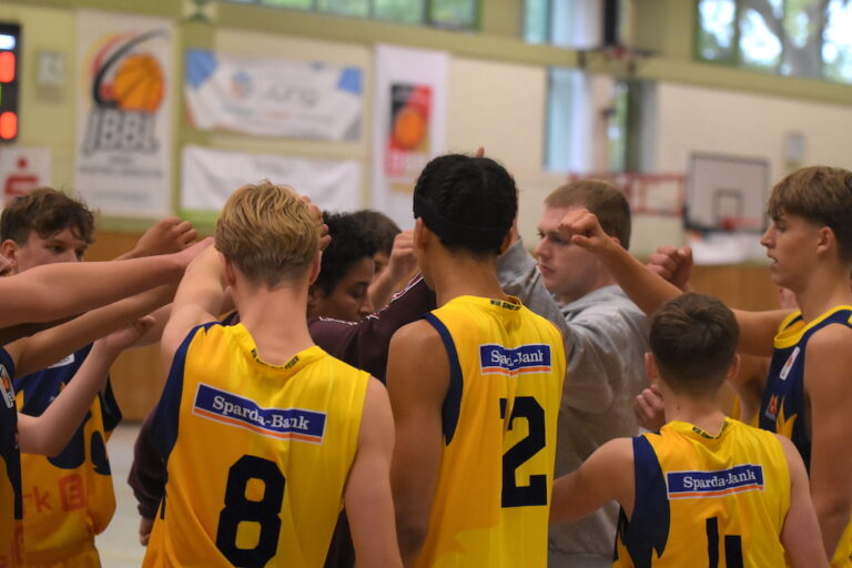 Mehrere junge Basketballspieler in gelben Trikots versammeln sich motiviert huddleartig zusammen.