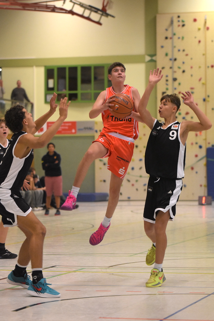 Ein jugendlicher Basketballspieler im orangefarbenen Trikot springt entschlossen zum Korb, flankiert von zwei Gegenspielern in Schwarz