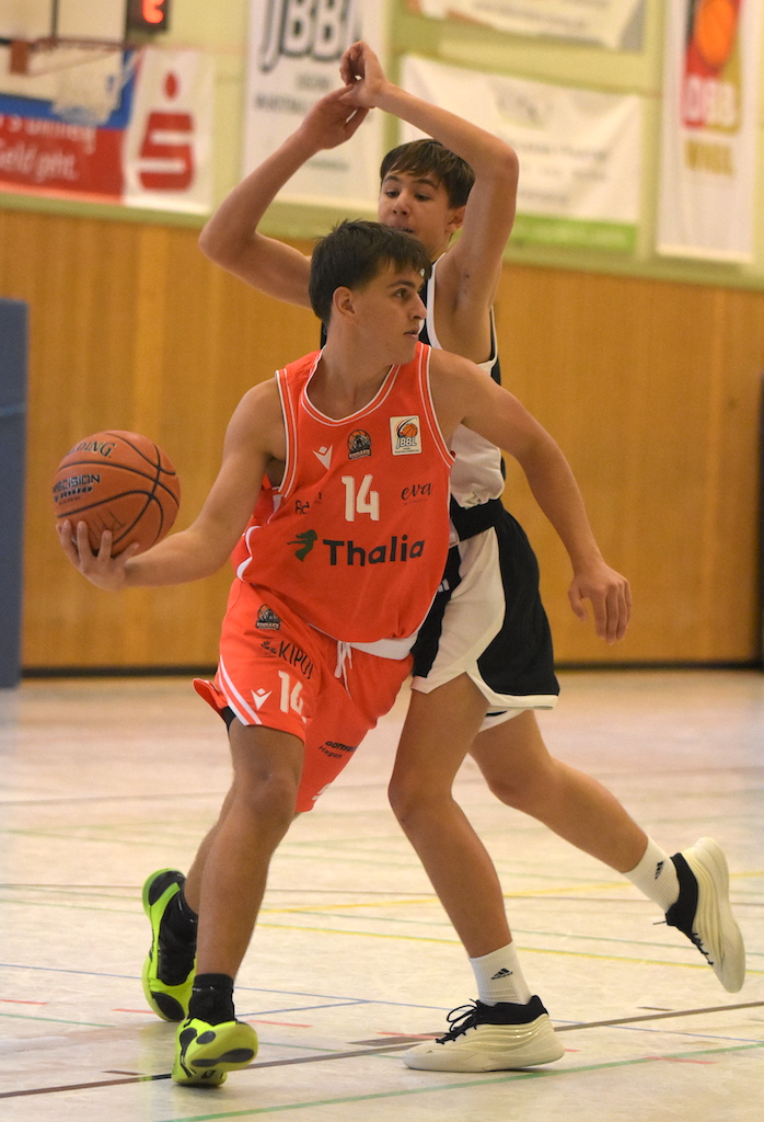 Ein junger Basketballspieler im orangenen Trikot mit ernster Miene wird verteidigt