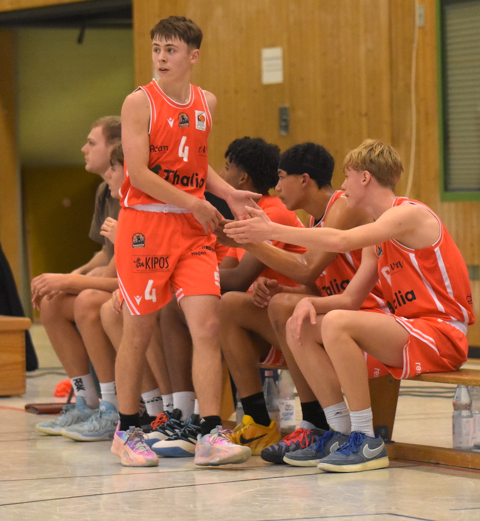Sechs junge Basketballspieler im orangefarbenen Trikot sitzen konzentriert auf einer Bank