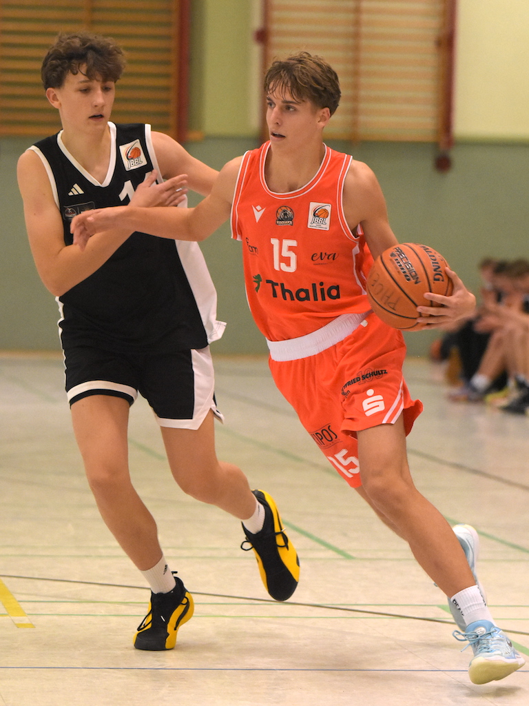 Zwei junge Männer spielen Basketball, einer in Orange mit Ball, der andere verteidigt energisch