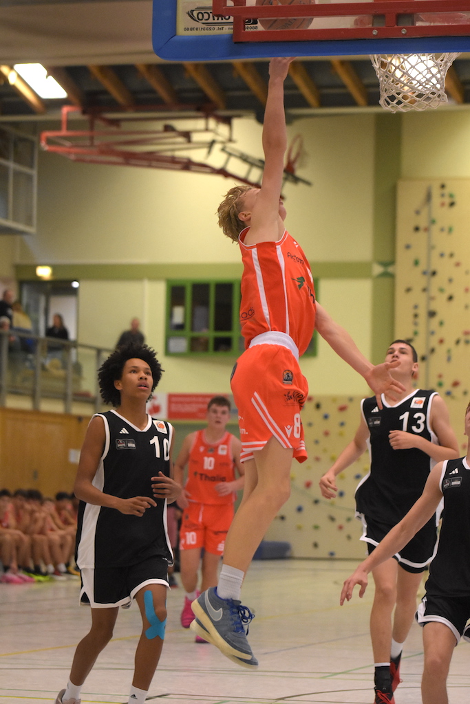Ein junger Basketballspieler im orangefarbenen Trikot dunkt energiegeladen am Korb