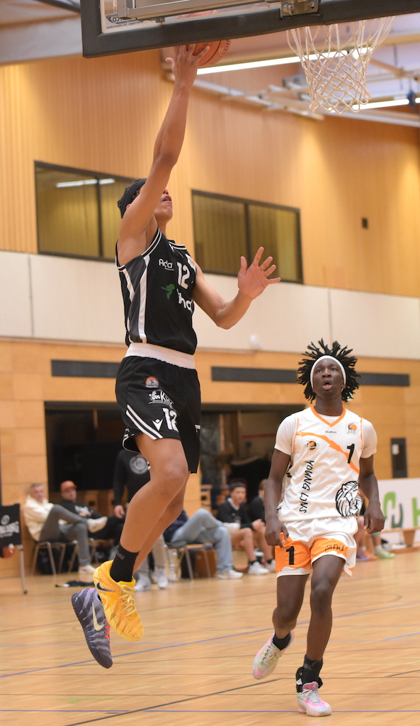 Ein Basketballspieler im schwarzen Trikot springt zum Korbleger, während ein Gegner zuschaut