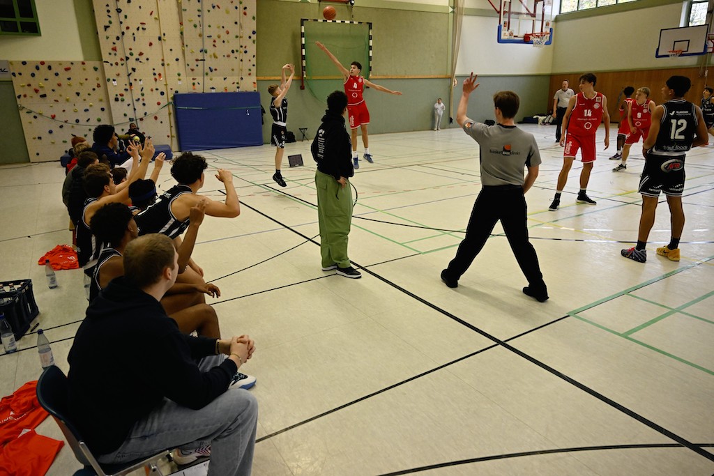 Ein Basketballspieler im schwarzen Trikot wirft einen Ball, während Mannschaftskameraden und Gegner aufmerksam zuschauen