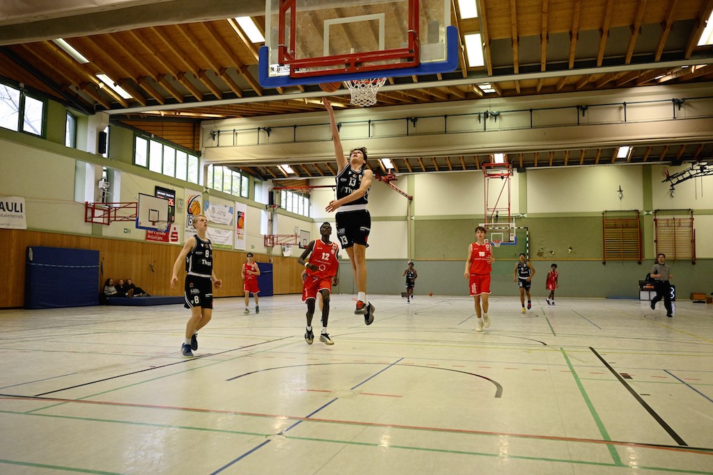 Ein Basketballspieler im schwarzen Trikot springt dynamisch zum Korb, Gegner in Rot beobachten energisch
