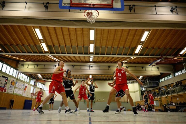 Mehrere Basketballspieler in roten und schwarzen Trikots schauen gespannt zum Korb