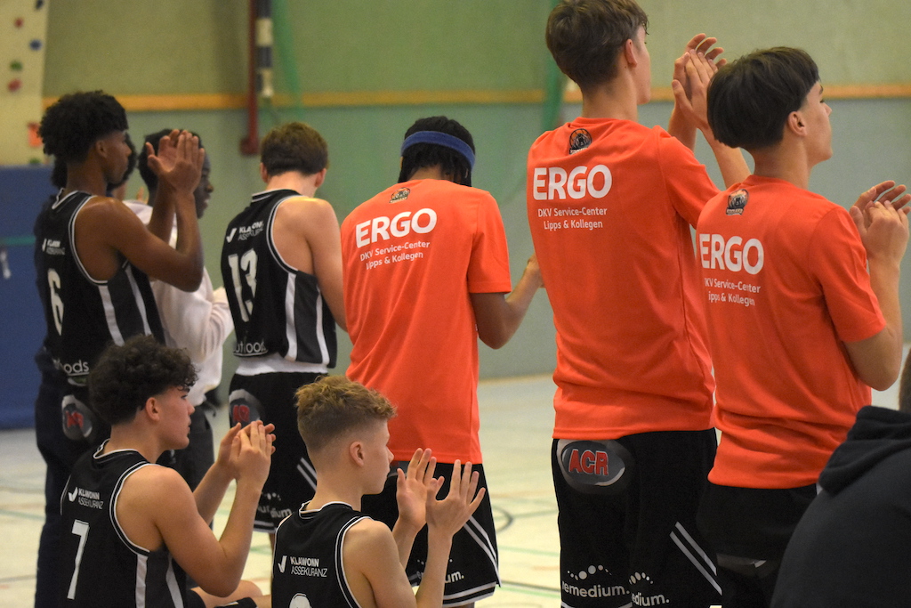 Mehrere junge Basketballspieler im Schwarz und Betreuer in Orange klatschen und jubeln