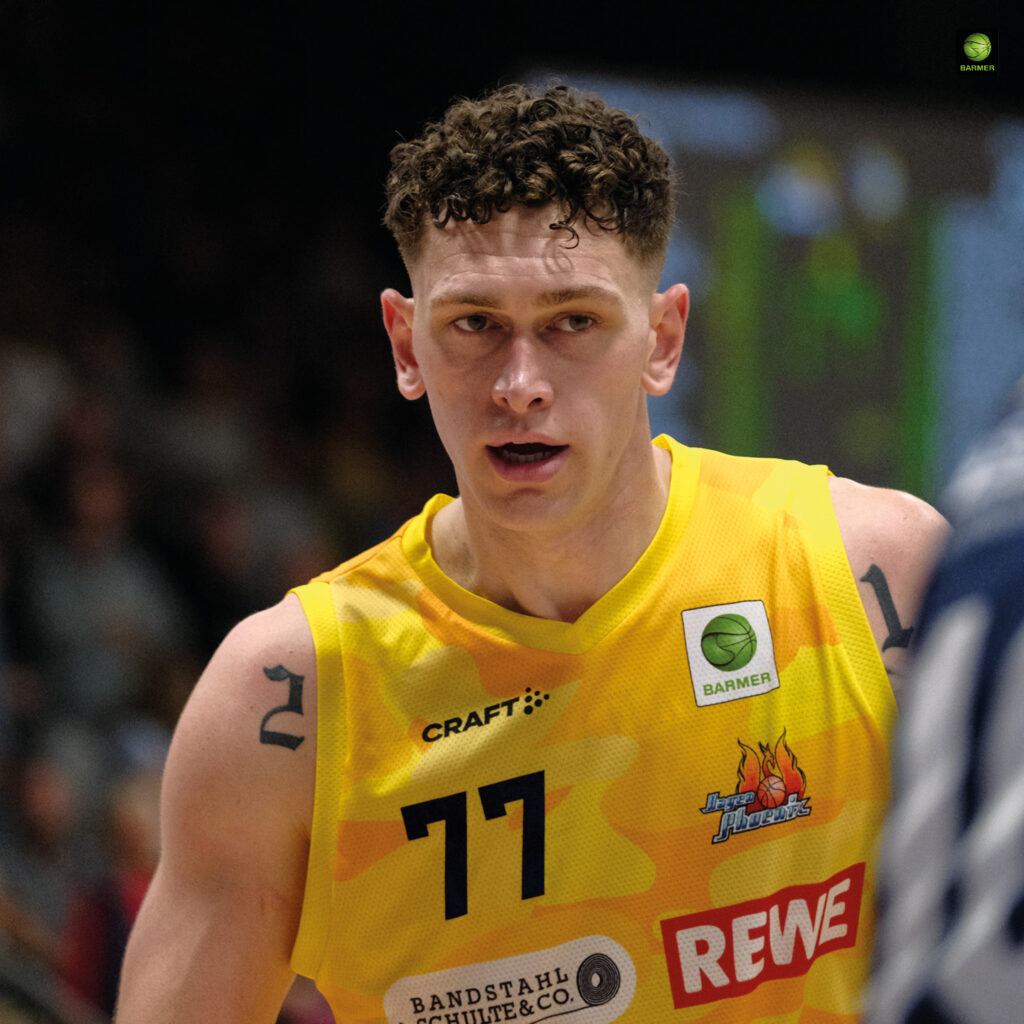 Ein junger Basketballspieler mit ernster Miene trägt ein gelbes Trikot und hat lockige Haare