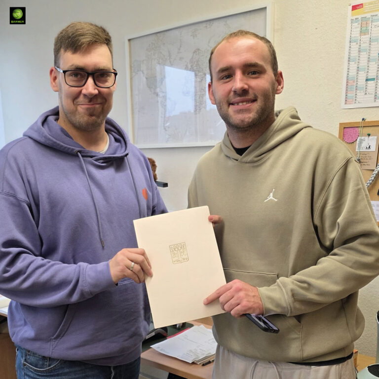 Zwei lächelnde Männer in beigen und lila Hoodies zeigen gemeinsam ein Blatt Papier
