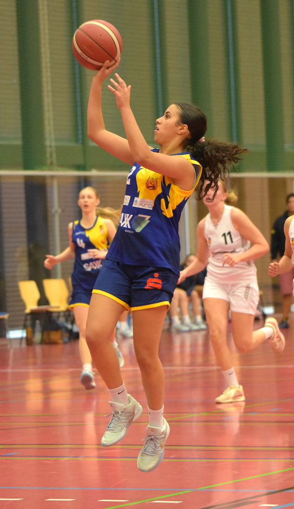 Eine junge Frau im blauen Basketballtrikot springt konzentriert zum Korbleger