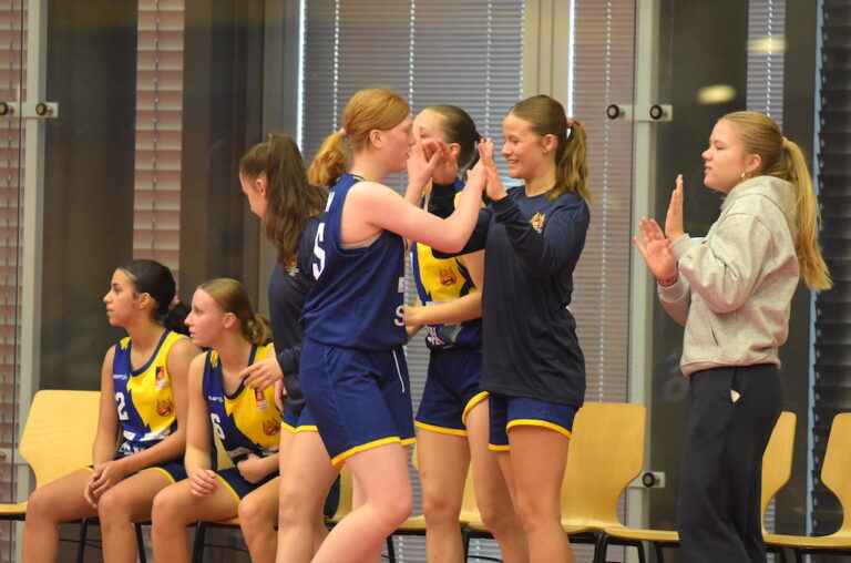 Junge Frauen im blauen Basketballtrikot jubeln lachend auf der Bank mit High Fives