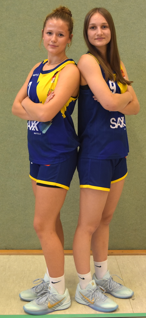Zwei junge Frauen in blauen Volleyballtrikots stehen Rücken an Rücken und lächeln