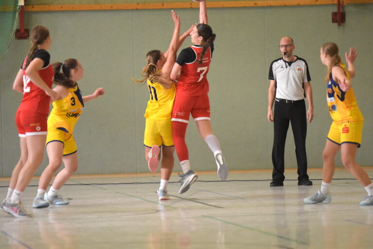 Eine Basketballspielerin im roten Trikot springt zum Korb, andere sehen zu