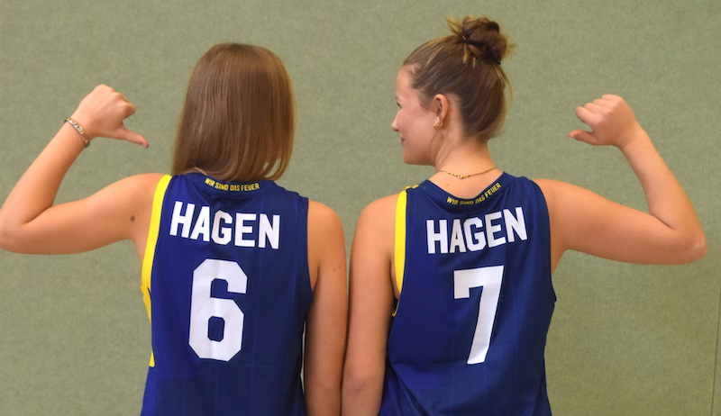 Zwei junge Frauen im blauen Trikot mit gelber Schrift zeigen lächelnd auf ihre Namen