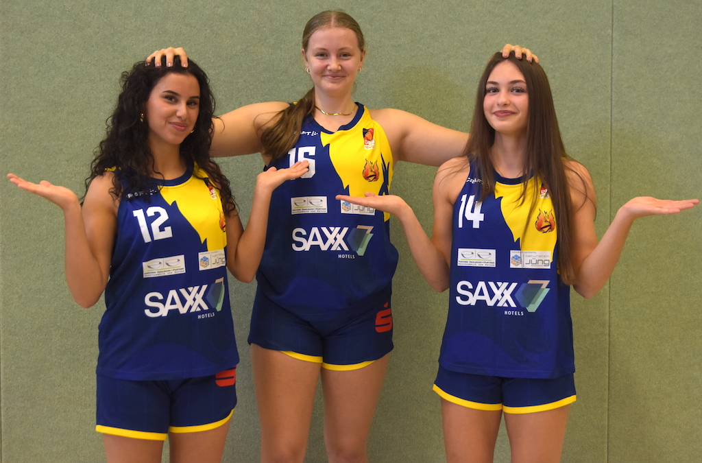 Drei junge Frauen im blauen Volleyballtrikot posieren lachend vor grüner Wand