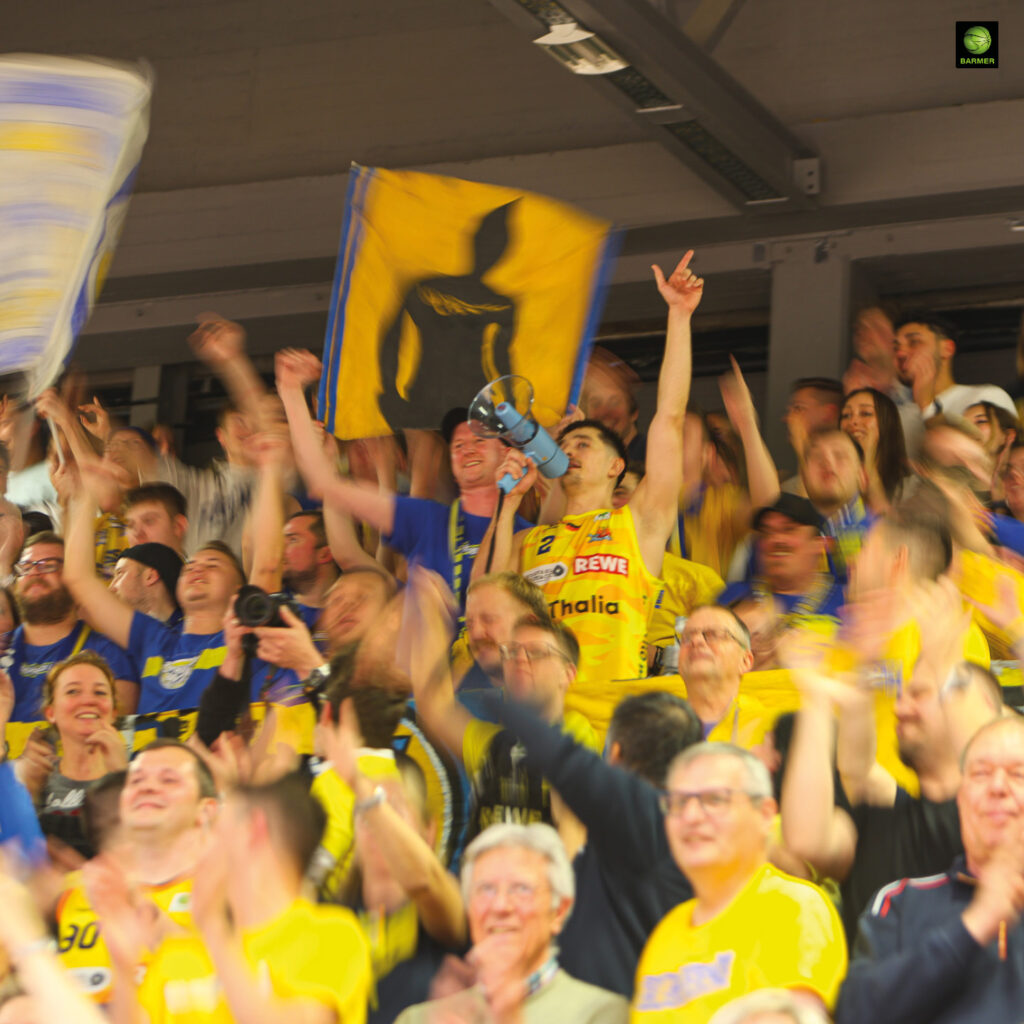 Jubelnde Männer in gelben Trikots halten Fahnen und zeigen freudige Gesichter