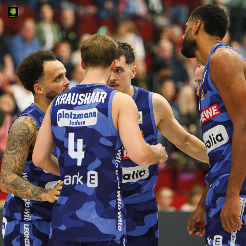 Vier Basketballspieler im blauen Trikot motivieren sich mit ernsten Mienen im Kreis