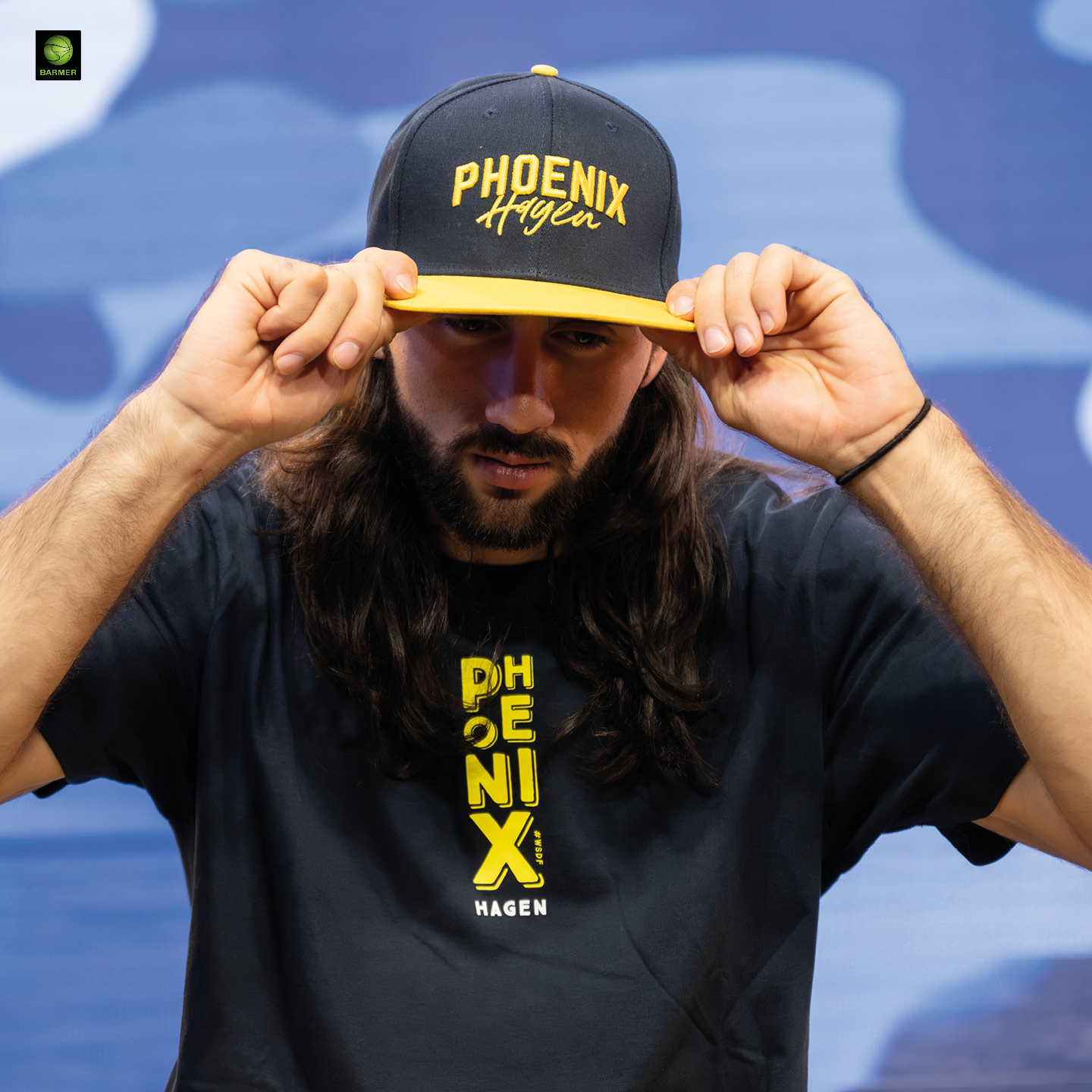 Ein Mann mit langem braunem Haar und Bart trägt ein Phoenix Hagen Shirt und Cap