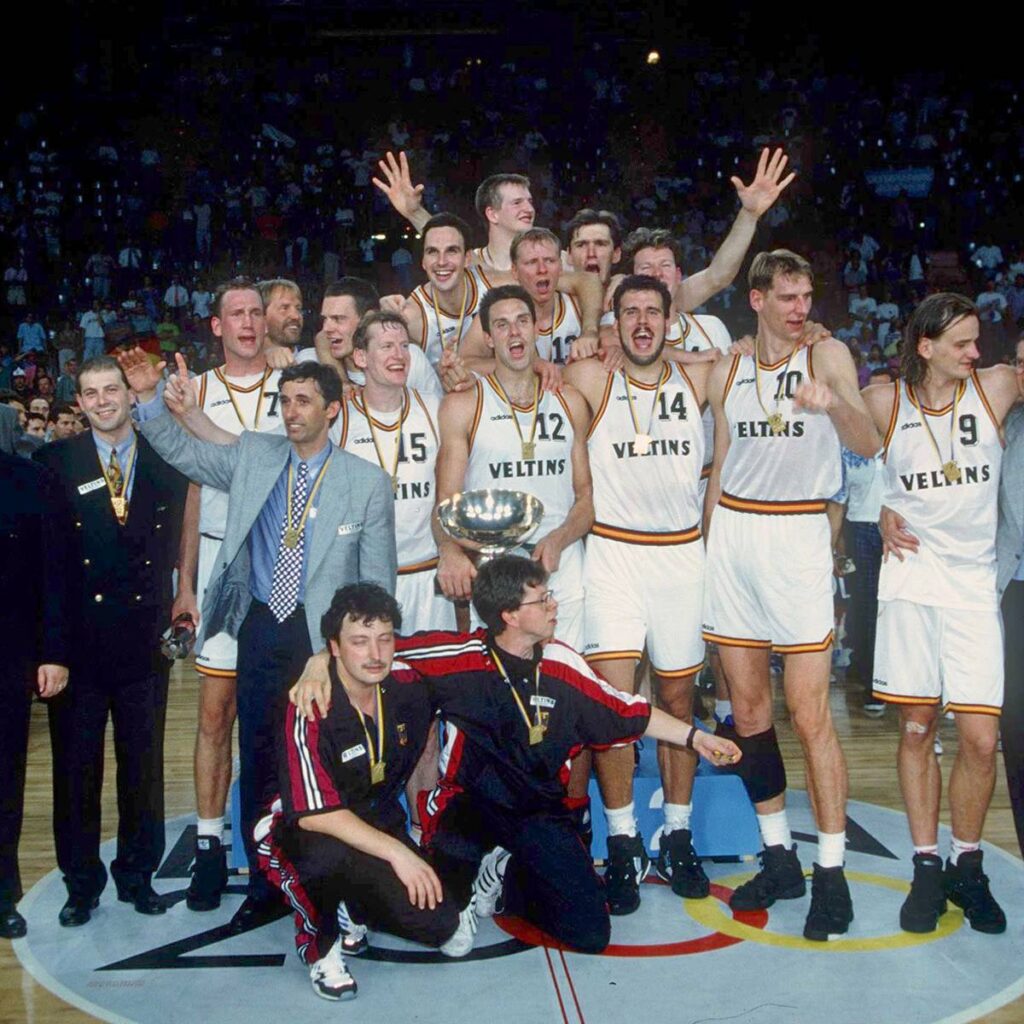 Eine jubelnde Basketballmannschaft in weißen Trikots feiert mit einem silbernen Pokal